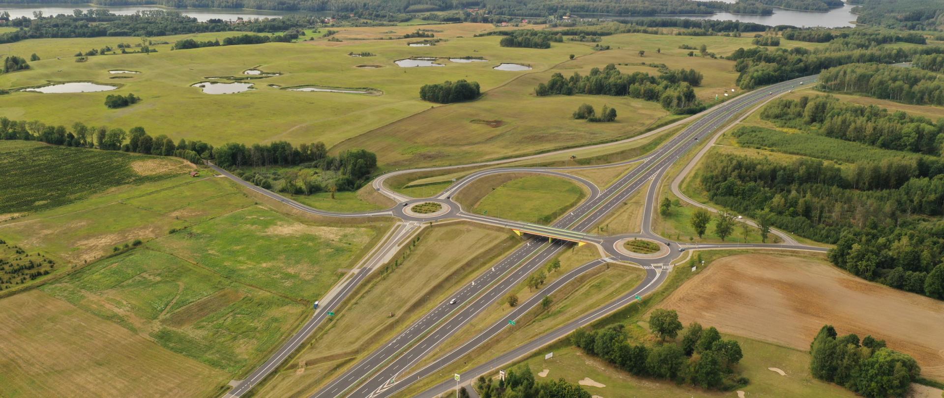 Widok z lotu ptaka. Na pierwszym planie dwupoziomowe skrzyżowanie, tzw. węzeł drogowy. Otoczenie drogi stanowią łąki, lasy i jeziora.