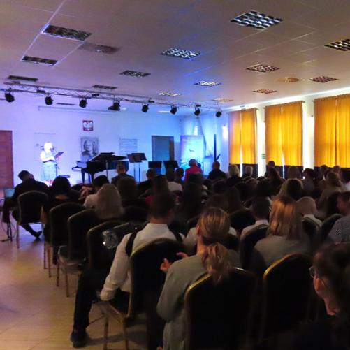 Zdjęcie przedstawia wypełnioną publicznością salę koncertową Państwowej Szkoły Muzycznej I stopnia im. J. S. Bacha w Grajewie. Po prawej stronie żółto-niebieskie zasłony, w tle scena. 