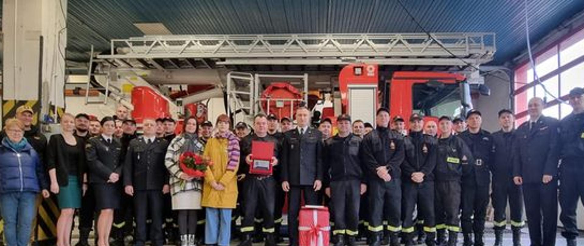 Na zdjęciu znajdują się strażacy oraz zaproszenie goście ustawieni w garażu na tle samochodu strażackiego