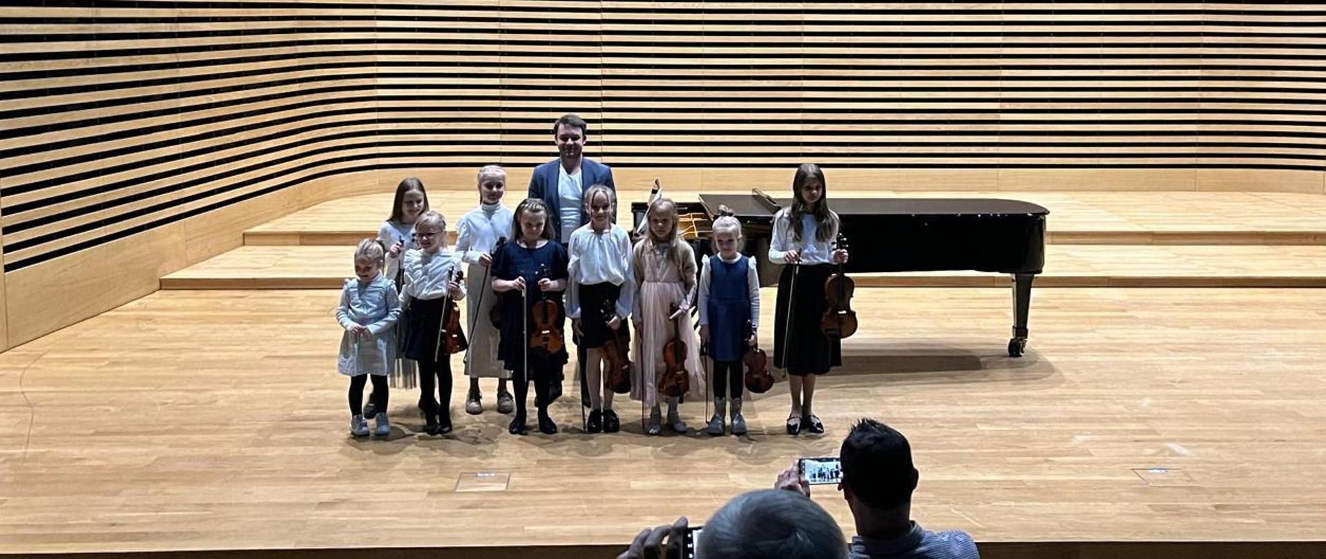 Dziewięć dziewczynek stoi w rzędzie i trzyma skrzypce, za nimi stoi mężczyzna.