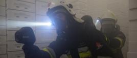 Widok z przodu. Rota gaśnicza OSP w trakcie przeszukania pomieszczeń. Strażacy korzystają z kamery termowizyjnej oraz latarek nahełmowych. W rękach trzymają linię gaśniczą zakończoną prądownicą. 