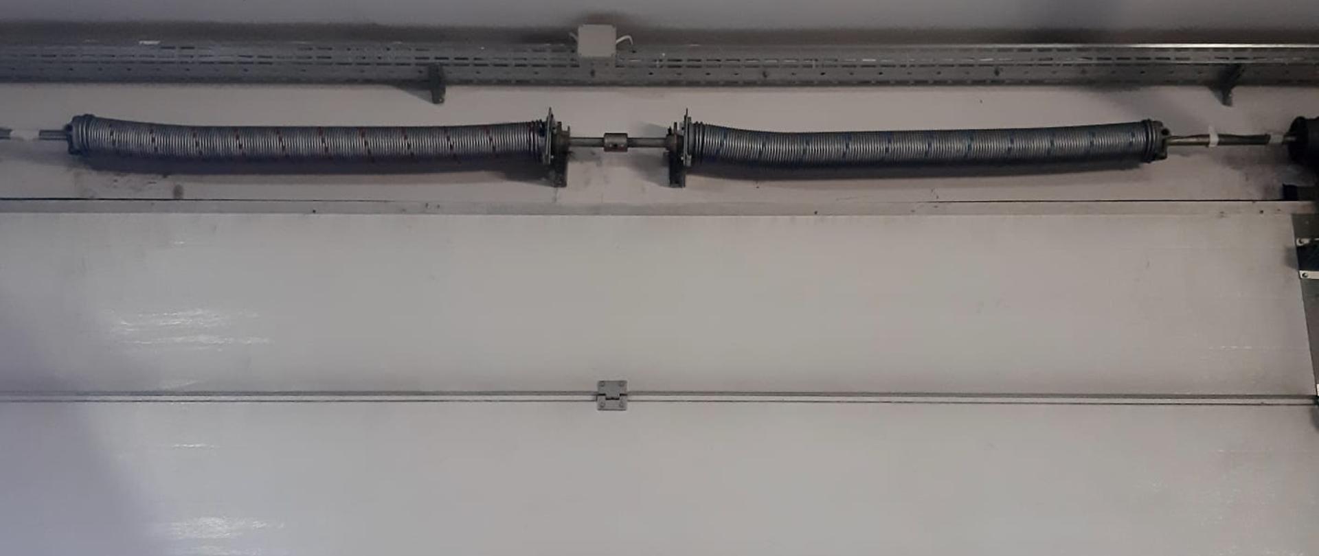 Na zdjęciu widzimy część bramy garażowej, panelowej koloru białego. Widzimy górną część bramy, dwa panele oraz komplet sprężyn stanowiących element mechanizmu podnoszenia bramy. Powyżej sprężyn uwidocznione jest także korytko z przewodami elektrycznymi. W dolnej części zdjęcia widzimy fragment trzeciego panelu bramy, który jest przeszklony i posiada czerwone ramy.