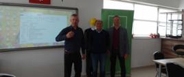 "Zdjęcie przedstawia trzech uśmiechniętych mężczyzn stojących obok siebie w jasnej sali konferencyjnej lub lekcyjnej.
Mężczyźni stoją w rzędzie na środku pomieszczenia:
• Mężczyzna po lewej ma krótkie, ciemne włosy i jest ubrany w ciemnoniebieską koszulę z długim rękawem oraz ciemne spodnie. Podnosi prawą dłoń na wysokości klatki piersiowej w gestzie przemawiania lub wskazywania.
• Mężczyzna w środku nosi okulary, ma krótkie włosy i szeroko się uśmiecha. Jest ubrany w jasnoniebieską koszulę z kołnierzykiem, ciemny sweter (v-neck) oraz klasyczne, niebieskie jeansy.
• Mężczyzna po prawej ma krótkie, jasne włosy i jest ubrany w bordowy t-shirt lub cienki sweter, szarą marynarkę oraz ciemne spodnie. Stoi wyprostowany z rękami opuszczonymi wzdłuż ciała.
Detale otoczenia:
• Po lewej stronie: Na ścianie zawieszona jest duża, włączona tablica interaktywna (lub ekran projekcyjny), na której wyświetlone jest okno systemowego menedżera plików. Bezpośrednio nad ekranem wisi godło Polski – biały orzeł w koronie na czerwonym tle. Pod sufitem zamontowany jest biały projektor multimedialny.
• W tle (za mężczyznami): Stoi zielony baner reklamowy (roll-up) z widocznym fragmentem adresu strony internetowej kończącym się na „.pl/k”. Tuż obok baneru, za środkowym mężczyzną, dostrzec można manekina ubranego w ciemną odzież ochronną i żółty kask budowlany.
• Po prawej stronie: Znajdują się duże okna wpuszczające naturalne światło, pod którymi na parapecie stoją małe, czerwone doniczki z kwiatami. Widoczny jest także fragment umeblowania sali – jasne stoły, nowoczesne czarne krzesła biurowe oraz drewniana lada w głębi. Podłoga wyłożona jest jasnymi kafelkami."