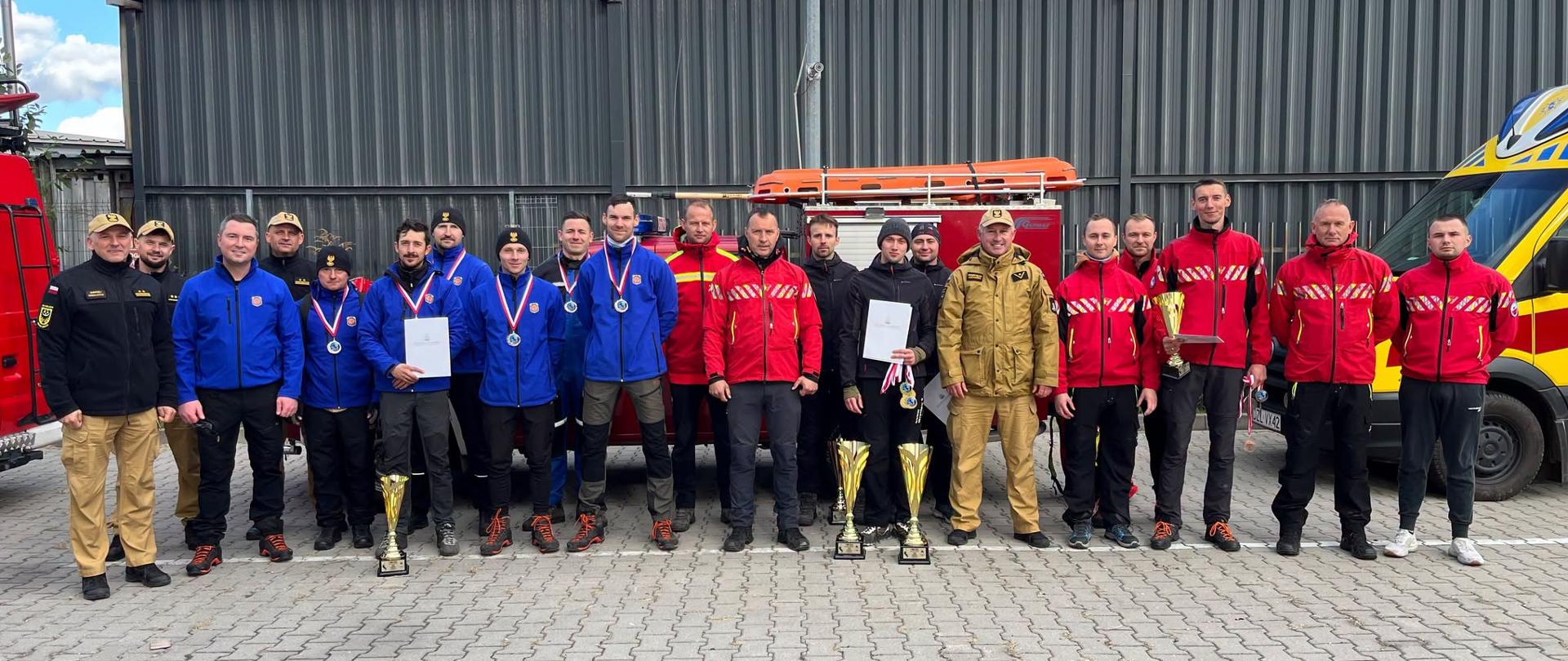 Grupowy portret kilkunastu mężczyzn – strażaków oraz ratowników – stojących w jednym rzędzie na wybrukowanym placu przed szarym, blaszanym budynkiem. Mężczyźni są ubrani w mundury i stroje służbowe w kolorach niebieskim, czerwonym, beżowym i czarnym. Wielu z nich ma na szyjach zawieszone medale, a niektórzy trzymają pamiątkowe dyplomy i złote puchary. Po lewej stronie widoczny jest fragment czerwonego wozu strażackiego, w tle stoi przyczepa ze sprzętem ratowniczym, a po prawej stronie znajduje się żółto-niebieski pojazd ratowniczy. Zdarzenie odbywa się w ciągu dnia, przy pogodnym niebie.