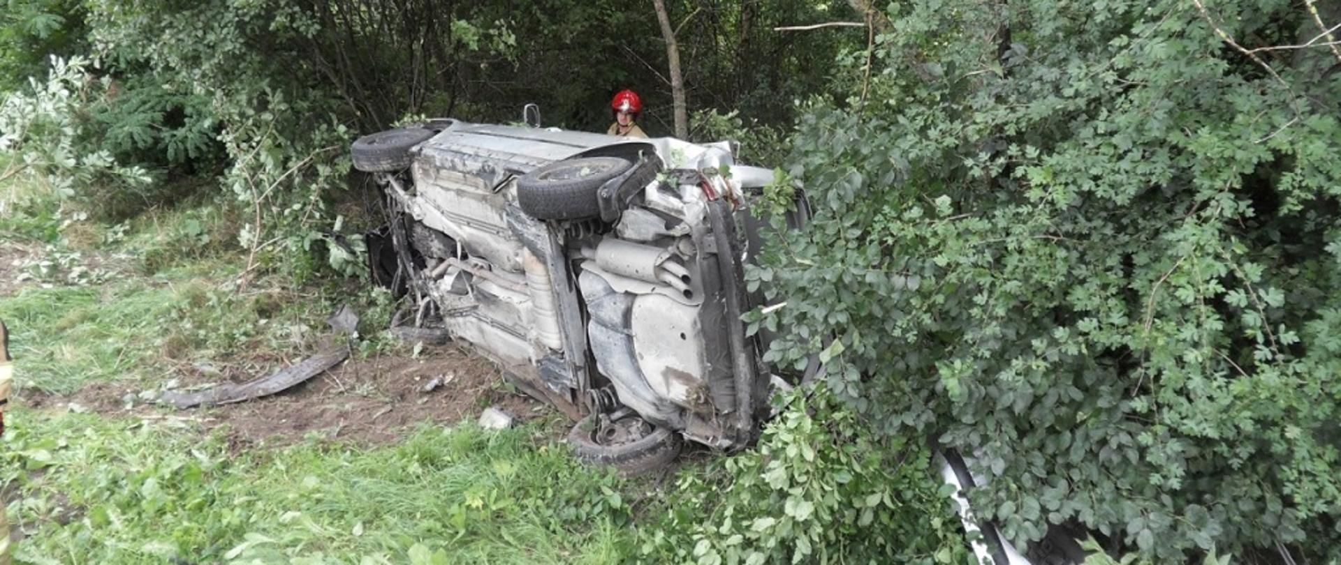 Zdjęcie przedstawia samochód osobowy leżący poza drogą na boku, za samochodem widać strażaka prowadzącego działania.