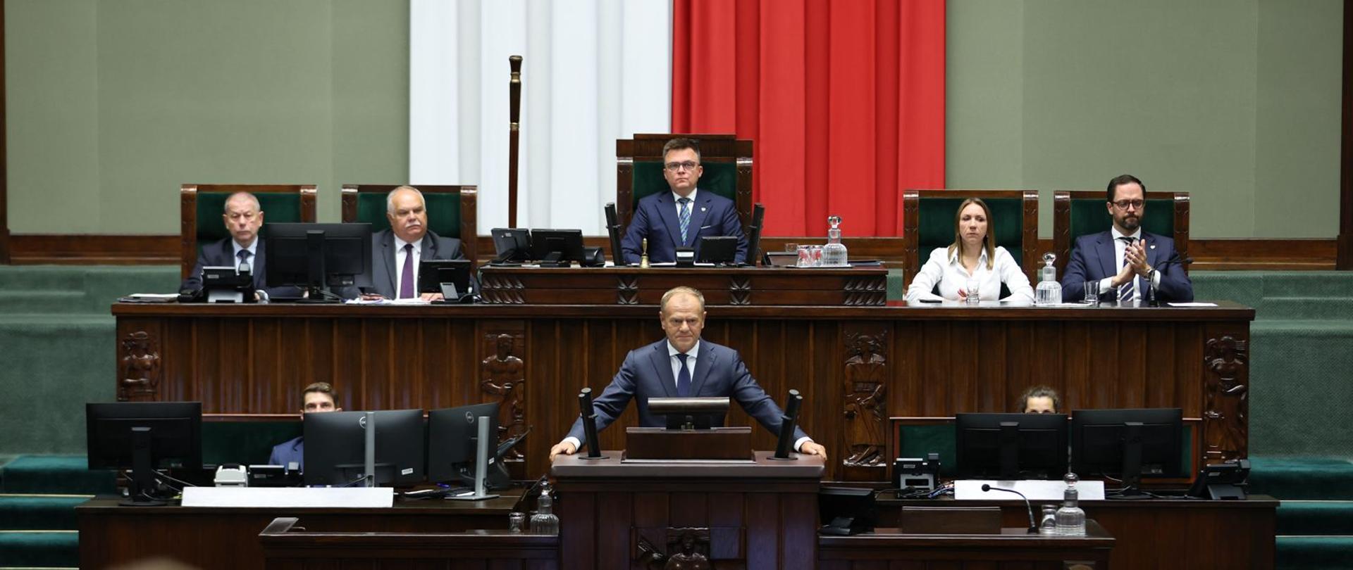 Premier Donald Tusk w trakcie wystąpienia w Sejmie. Stoi na mównicy. W tle Marszałek Sejmu Szymon Hołownia.