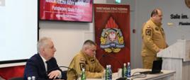 Strażak stoi przy mównicy i przemawia obok siedzą przy stole funkcjonariusz oraz mężczyzna.