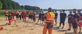 Zdjęcie przedstawia strażaków PSP oraz OSP podczas instruktażu podejmowania osób poszkodowanych z wody na plaży przy jeziorze głuszyńskim.