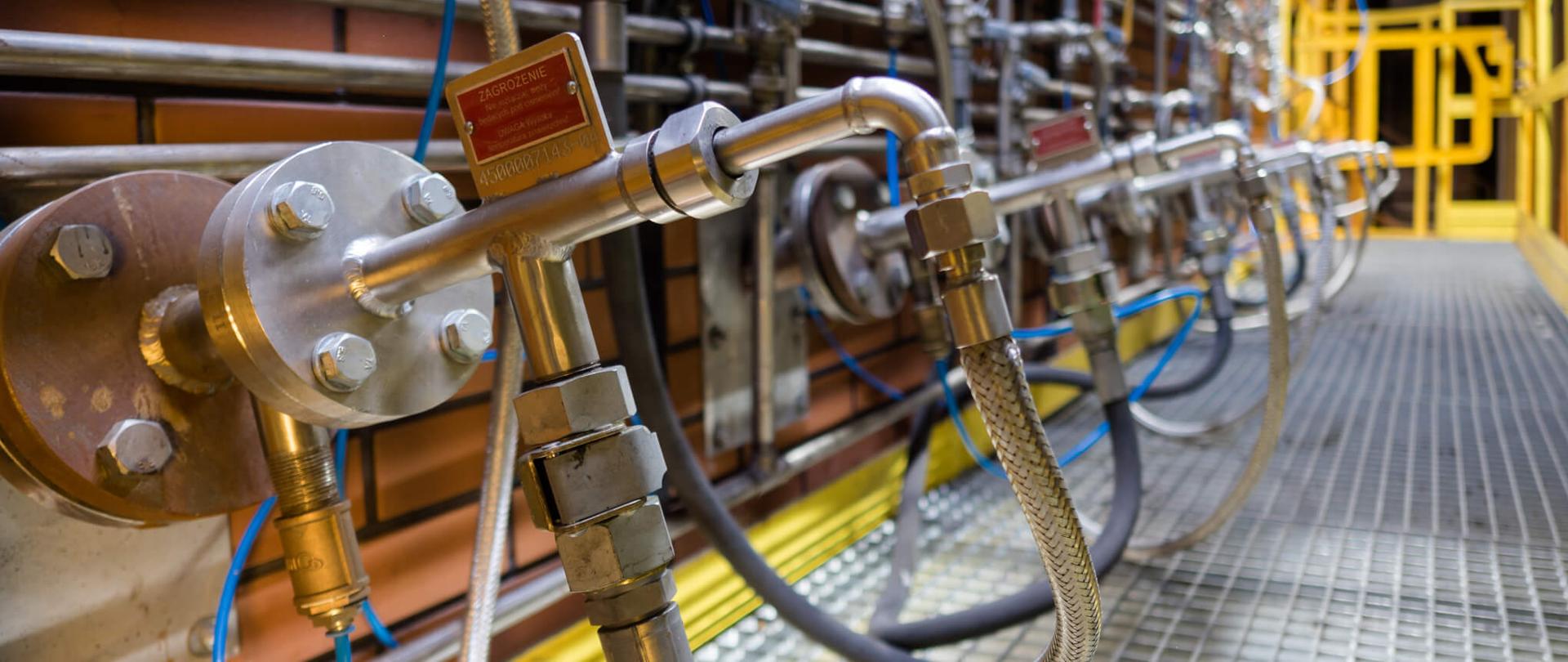 Instalacja na kotle. Moduł odazotowania spalin. Widoczna ściana, na której zamontowana jest konstrukcja z gumowymi i metalowymi przewodami oraz srebrne zawory. Podłoże stanowi metalowa kratka.