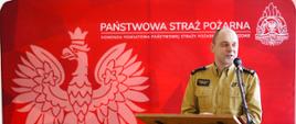 Przedstawiciel Wielkopolskiego Komendanta Wojewódzkiego Państwowej Straży Pożarnej w trakcie przemowy przed mównicą. W tle ścianka reklamowa komendy z godłem Polski i logiem Państwowej Straży Pożarnej.