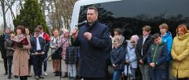 Minister Czarnek stoi na wyłożonym szarą kostką podwórku i mówi do mikrofonu na stojaku, za nim grupa dzieci stoi pod białym autokarem.