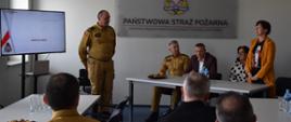 Na zdjęciu widoczna sala. Przy białych Stołach siedzą uczestnicy narady. Na ścianie w tle widoczne logo Państwowej Straży Pożarnej. Dwie z osób stoją i przemawiają do reszty.