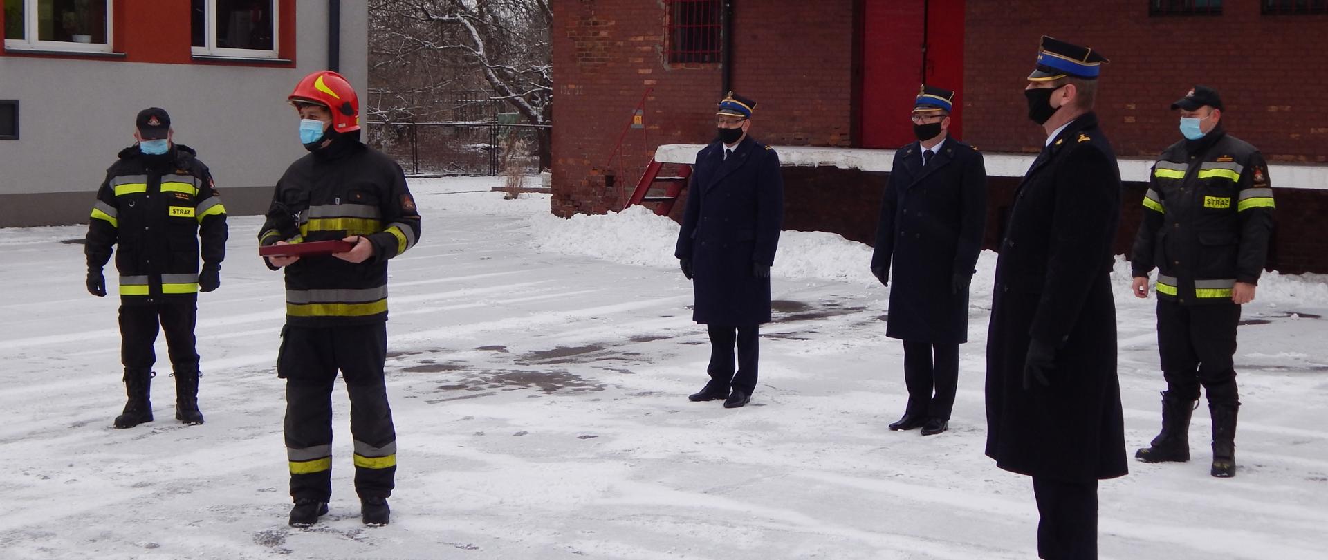 Zdjęcie przedstawia funkcjonariuszy Państwowej Straży Pożarnej podczas uroczystej zamiany służby. Na zdjęciu widać funkcjonariuszy w umundurowaniu wyjściowym oraz specjalnym na tle budynku Jednostki Ratowniczo Gaśniczej nr 1