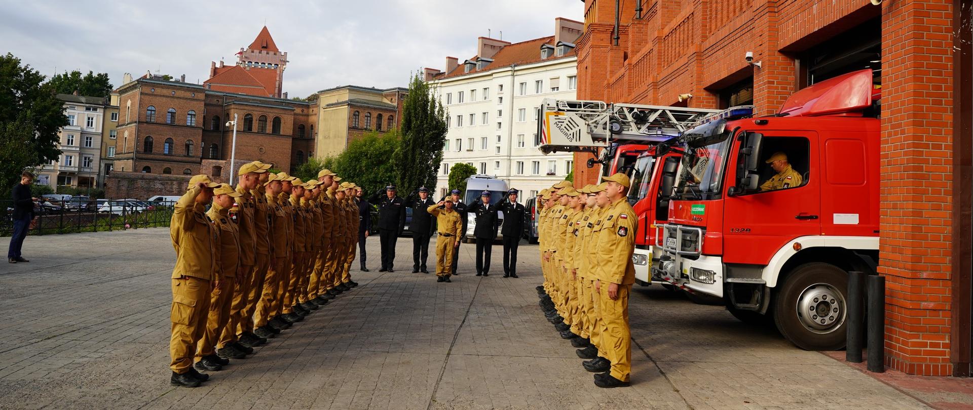 uroczysta zbiórka przed jednostką strażacką, w dwóch szeregach na przeciwko siebie stoją strażacy, w tle pojazdy pożarnicze