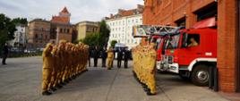 uroczysta zbiórka przed jednostką strażacką, w dwóch szeregach na przeciwko siebie stoją strażacy, w tle pojazdy pożarnicze