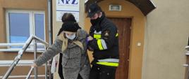 Zdjęcie przedstawia strażaka OSP, który trzyma pod ręką starszą kobietę i pomaga jej zejść ze schodów. W tle znajduje się budynek Niepublicznego Zakładu Opieki Zdrowotnej w Ostrowitem, gdzie znajduje się punkt szczepień. Kobieta oraz strażak mają założone maseczki chirurgiczne na twarzy. Na schodach i poza nimi widać leżący dookoła śnieg. 
