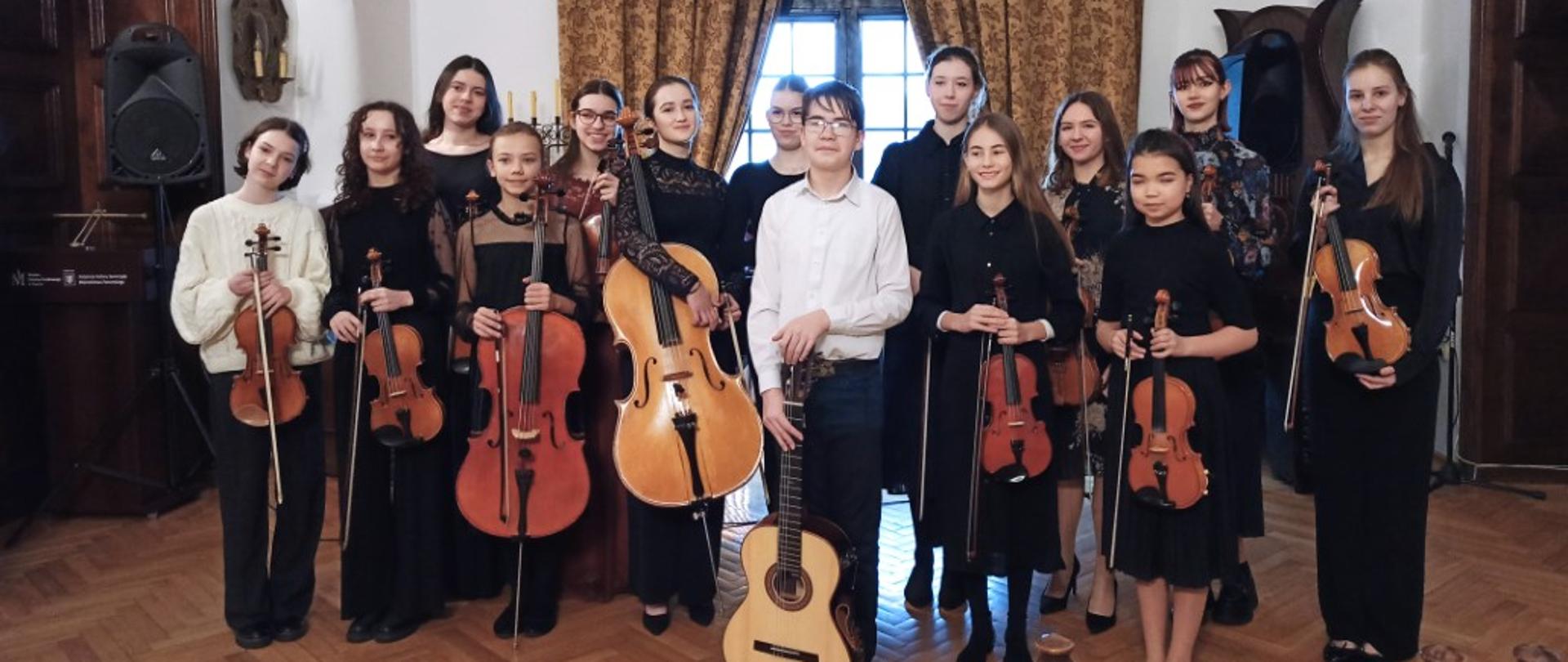 Zdjęcie kolorowe. Odświętnie ubrani uczniowie ze skrzypcami, wiolonczelami, gitarą pozują do zdjęcia. W tle okno z ozdobnie upiętymi zasłonami.