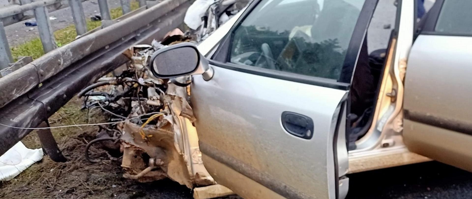 Zdjęcie przedstawia uszkodzony samochód osobowy po wypadku drogowym. Pojazd stoi na jezdni przed metalowymi barierami energochłonnymi