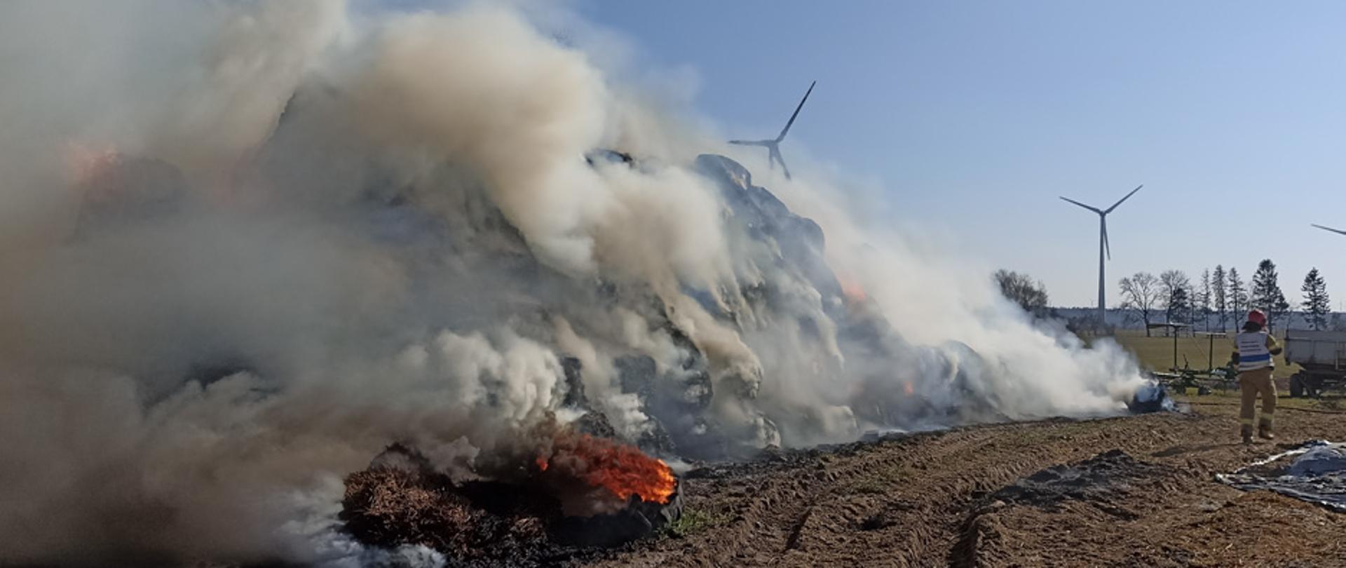 Gęsty dym unoszący się nad stertą balotów, obok strażak i przyczepa ciągnikowa