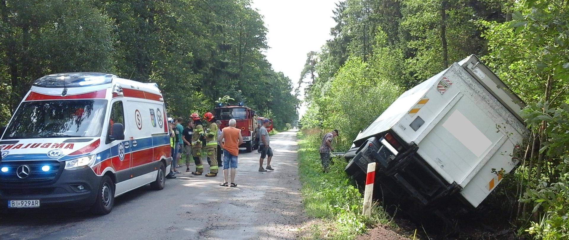 Samochód ciężarowy stoi przechylony w przydrożnym rowie. Na drodze stoi karetka, a za nią samochody ratowniczo-gaśnicze.