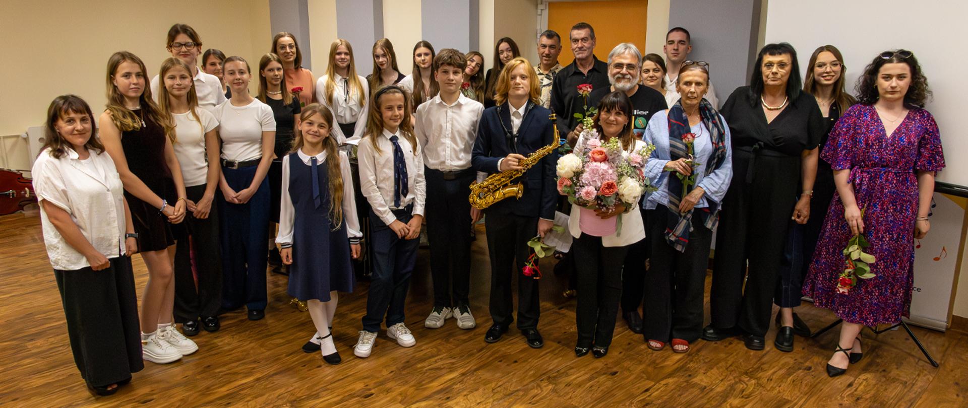 Dyrektor, nauczyciele i absolwenci stoją w auli szkolnej, pozują do zdjęcia.