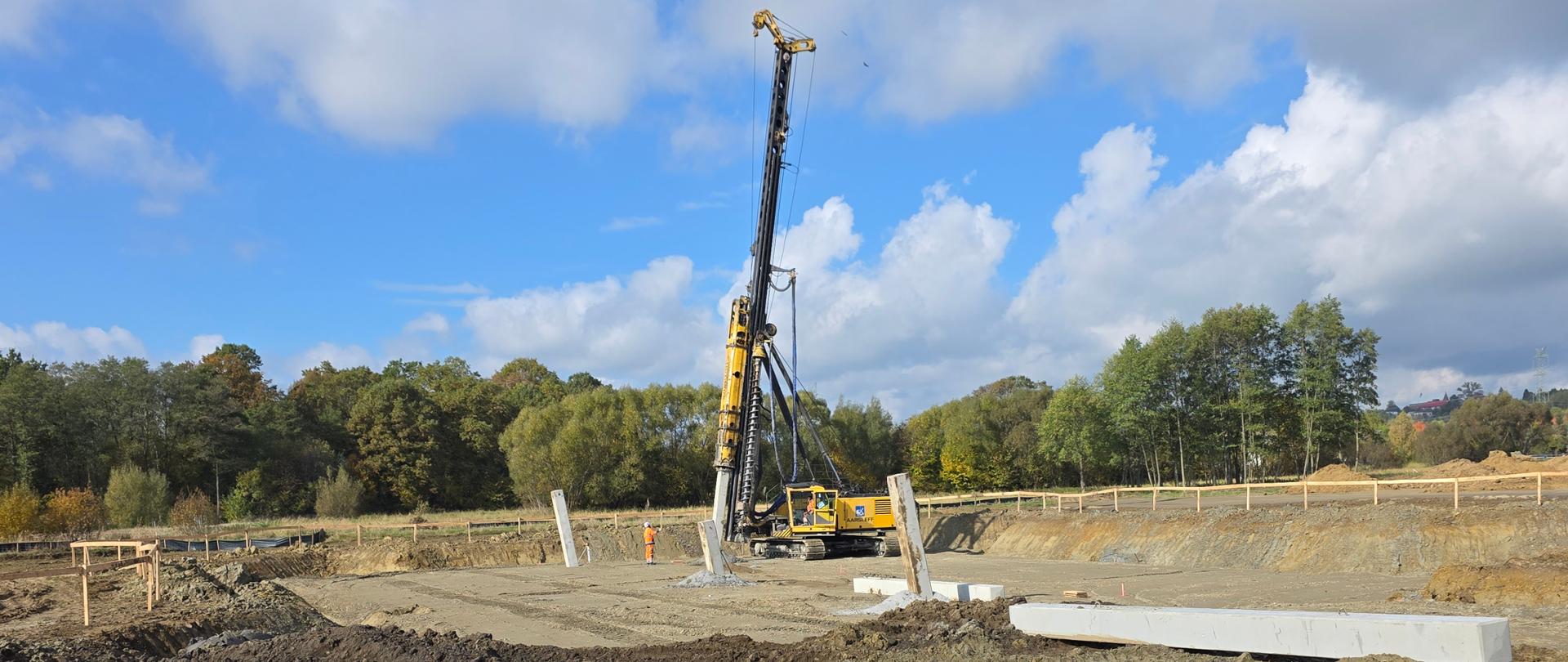 Zdjęcie przedstawia etap wykonywania próbnych pali na placu budowy. W centralnej części znajduje się duża maszyna palownicza (żółta), wyposażona w wysoki pionowy maszt, służąca do wbijania lub wiercenia pali w grunt. Obok maszyny widoczne są betonowe elementy – prawdopodobnie prefabrykowane pale lub belki. eren jest częściowo wyrównany, z widocznymi śladami prac ziemnych (wykopów i nasypów). W tle znajdują się drzewa w jesiennej kolorystyce, co sugeruje porę roku – jesień. Niebo jest częściowo zachmurzone, ale z przejaśnieniami, co daje dobre oświetlenie.