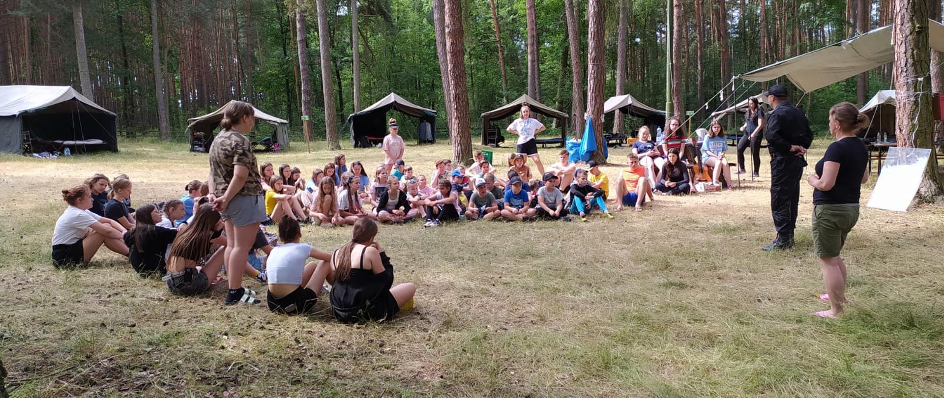 Zdjęcie przedstawia strażaka prowadzącego pogadankę na temat bezpiecznych zachowań dla uczestników obozu harcerskiego. Widoczny las a w tle, namiotu obozu w kolorze zielonym