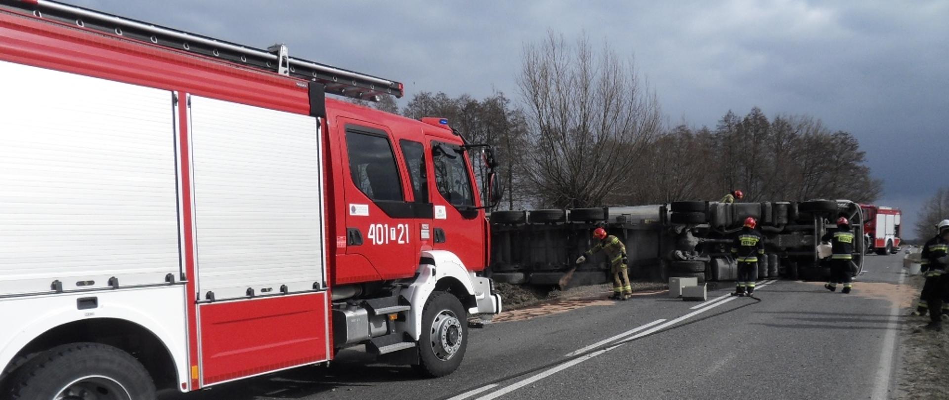 Na zdjęciu przewrócony pojazd ciężarowy, przy nim pracujący strażacy, po lewej stronie samochód pożarniczy o raz radiowóz policji