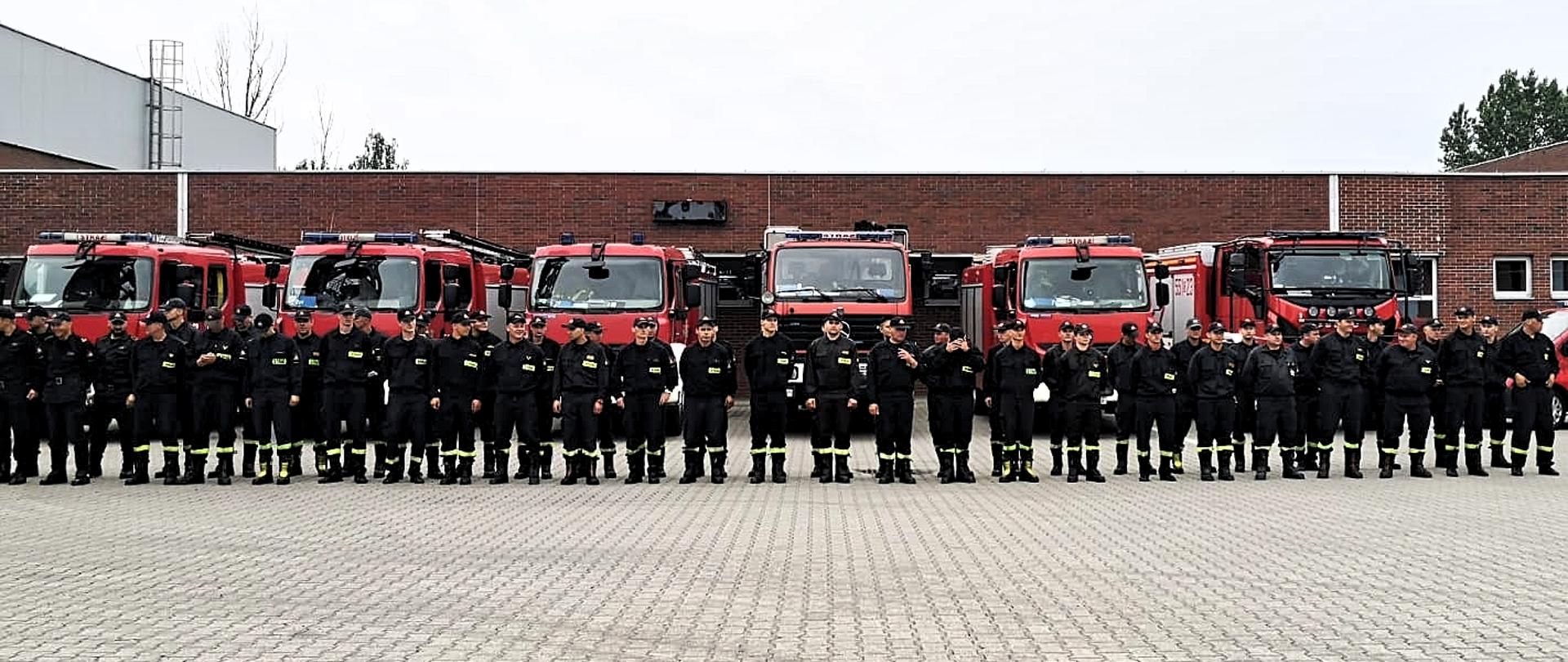 Strażacy ustanowieni w szeregu na tle wozów strażackich 