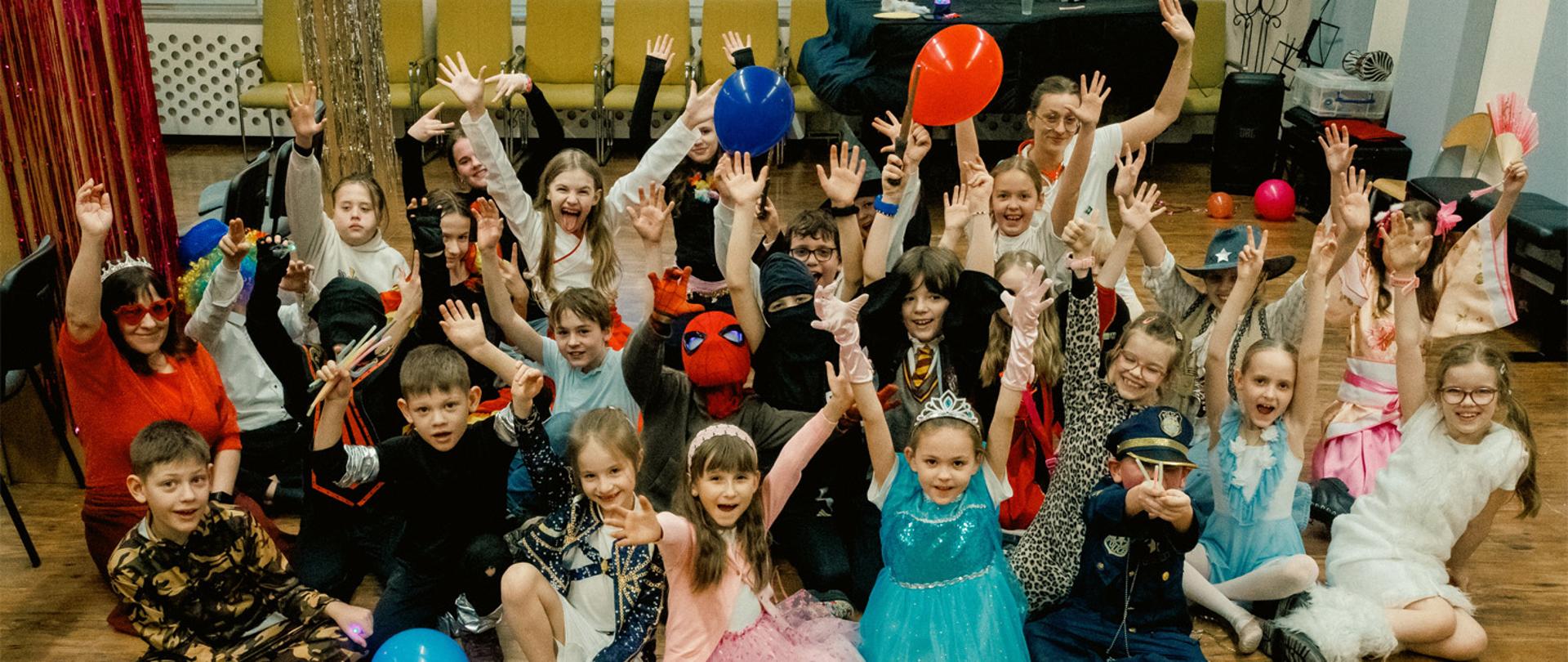 Grupa kilkudziesięciu dzieci siedzi na podłodze, wszyscy są przebrani w stroje karnawałowe, trzymają ręce w gorze.