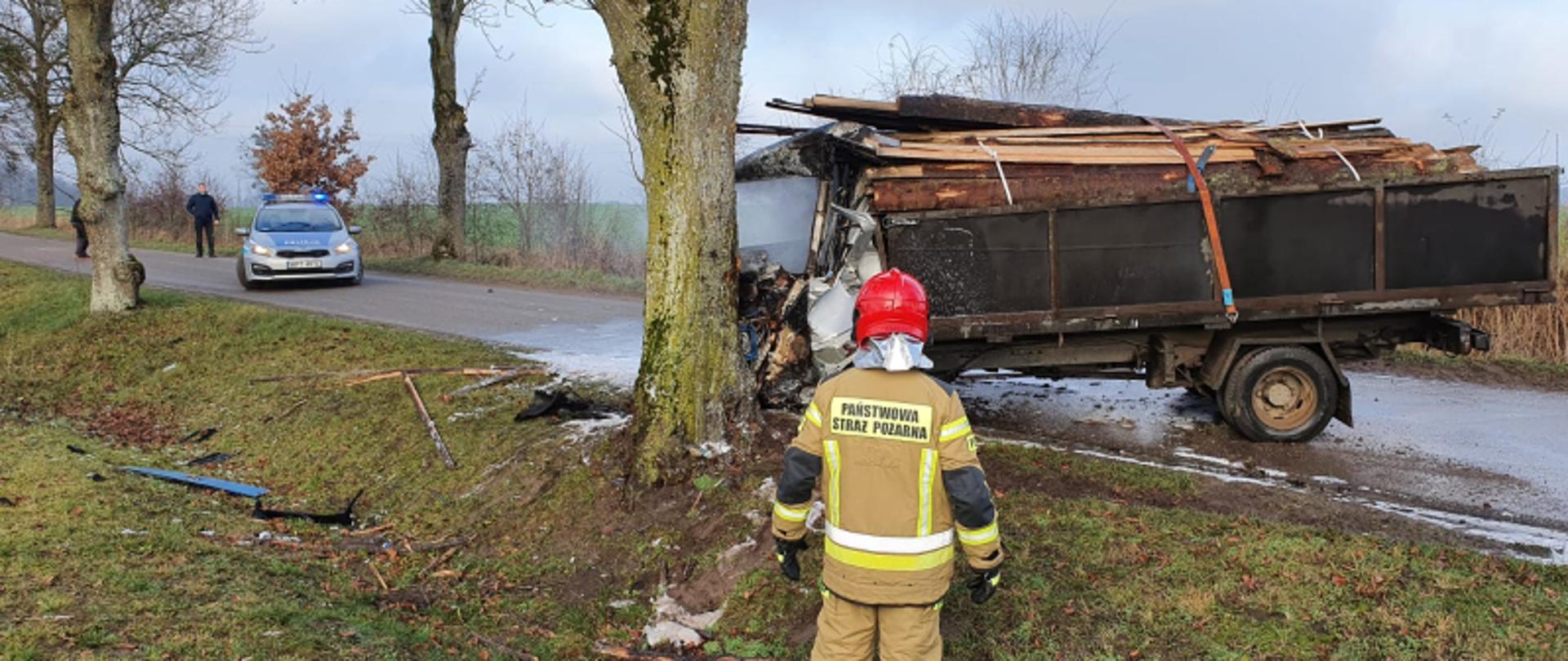 Na zdjęciu widoczny strażak stojący naprzeciw wraku ciężarówki. Pojazd po uderzeniu w drzewo z deformacją szoferki oraz spaloną kabiną. Za pojazdem znajduję się patrol policji.
