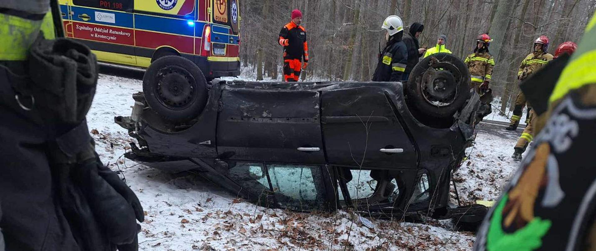 „Samochód osobowy leżący na dachu przy zaśnieżonej leśnej drodze; wokół strażacy i ratownicy, w tle karetka.”