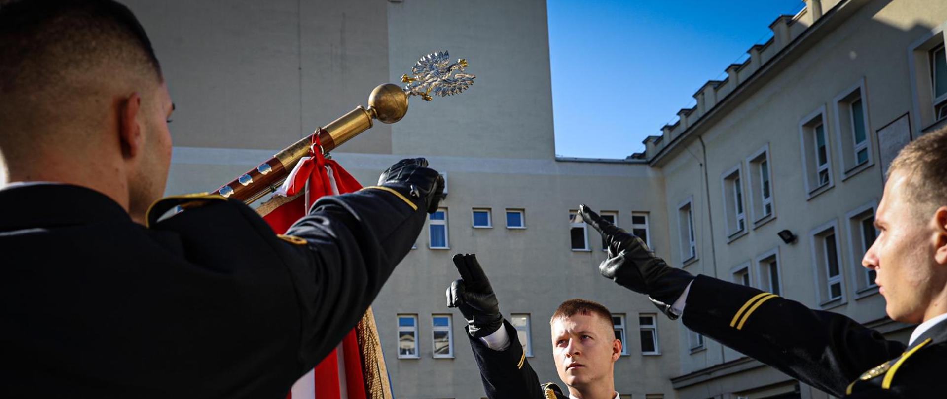 Ślubowanie na sztandar. Trzech słuchaczy składa rotę ślubowania. Mężczyźni – strażacy – w umundurowaniu wyjściowym z wyciągniętymi rękoma w stronę płachty sztandaru. 