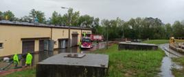 Strażacy wypompowują wodę z zalanego placu na terenie oczyszczalni ścieków. Po lewej budynki gospodarcze, w tym rozdzielnia prądu w kolorze piaskowym. Chodniki wzdłuż budynku całkowicie zalane. W rozlewisku stoją dwa pojazdy pożarnicze. Wzdłuż budynku rozciągnięte węże , którymi strażacy odprowadzają wodę z rozlewiska. Po prawej instalacje oczyszczalni. 