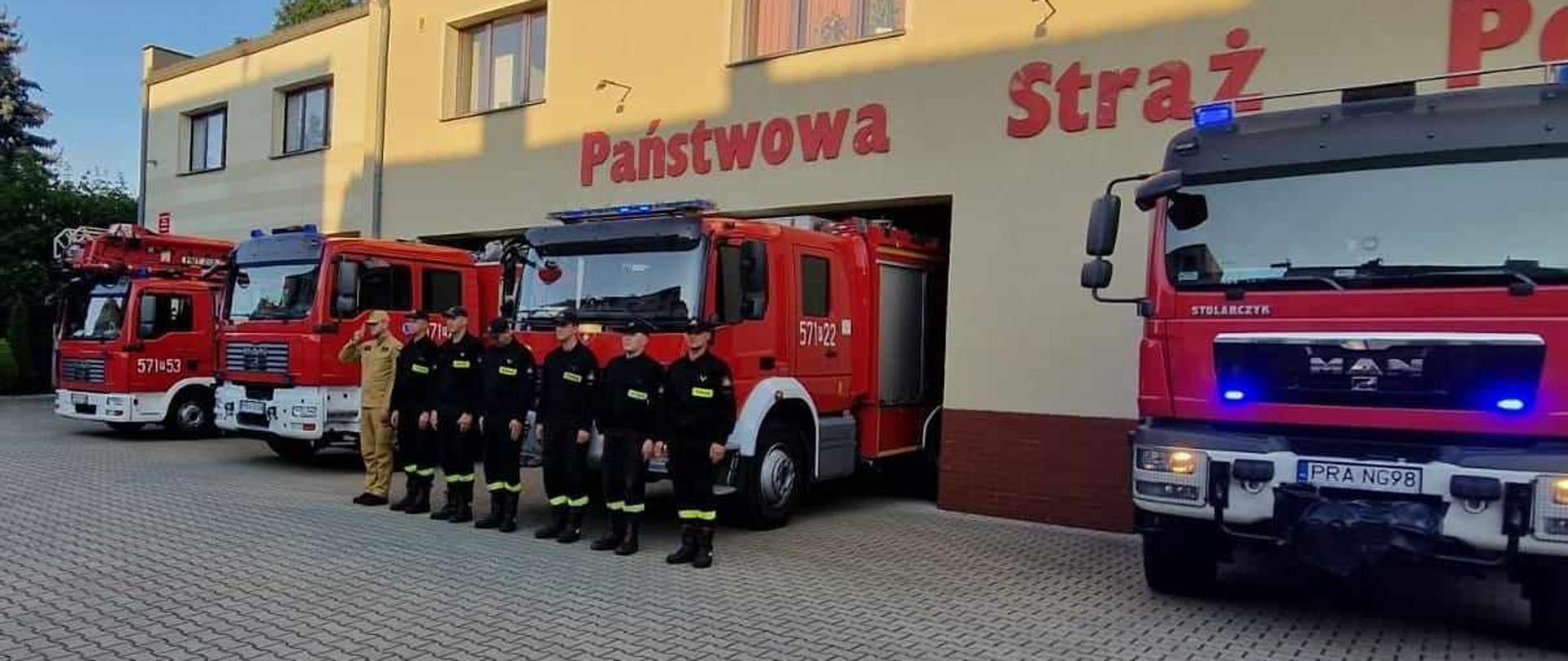 Uczczenie pamięci tragicznie zmarłego strażaka. Zdjęcie przedstawia budynek komendy powiatowej PSP w Rawiczu. Pomiędzy oknami a bramami garażowymi widnieje czerwony napis Państwowa Straż Pożarna. Przed bramami garażowymi stoją 4 pojazdy strażackie z włączonymi sygnałami świetlnymi.Przed jednym z wozów, w szeregu stoi 7 strażaków w ubraniach koszarowych. Dowódca zmiany oddaje honor poprzez salutowanie.