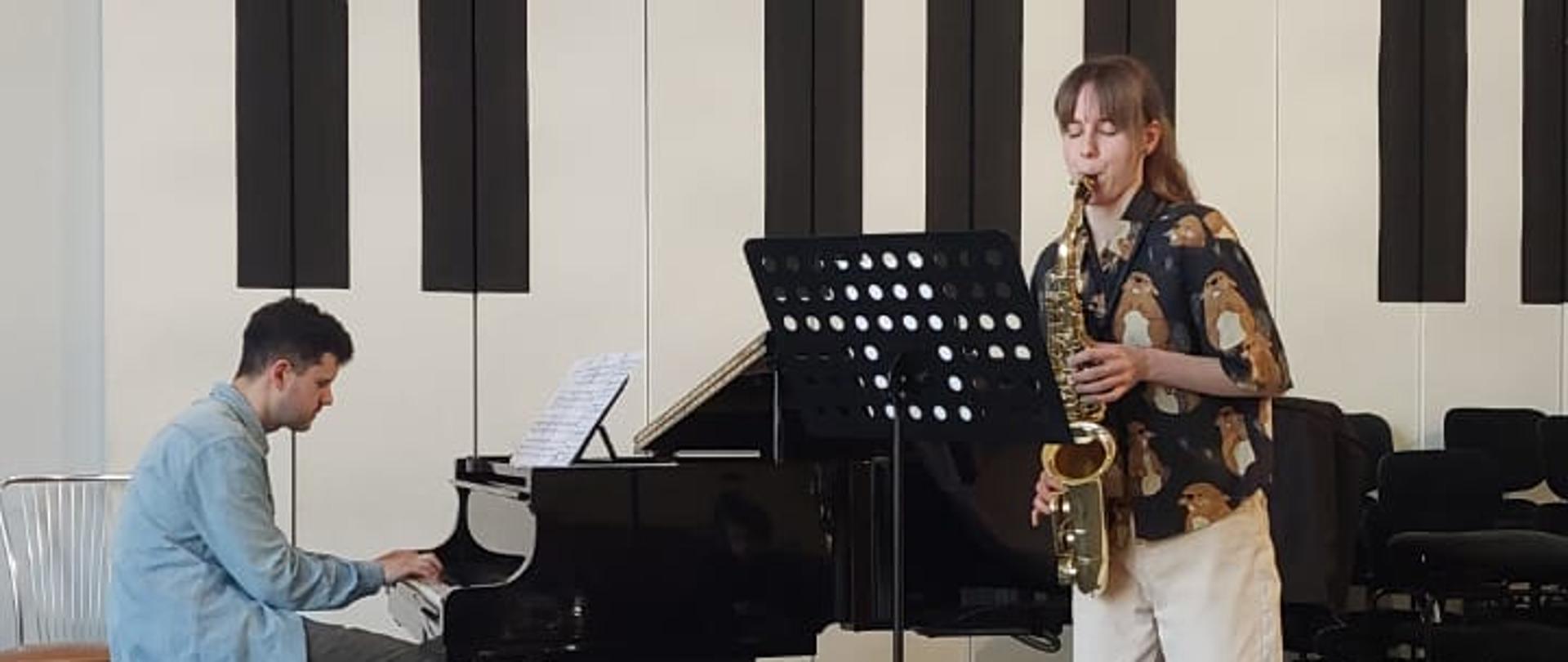 Dziewczyna gra na saksofonie, mężczyzna akompaniuje jej na fortepianie.