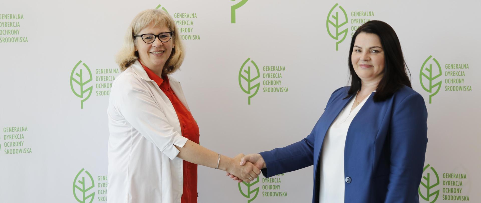 Na tle białej ściany z logotypem Generalnej Dyrekcji Ochrony Środowiska (zielony liść) stoją dwie uśmiechnięte kobiety. Ściskają sobie dłonie. 
