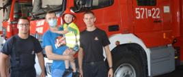 Wspieramy leczenie Gosi. Laureat licytacji "Dnia ze strażakami w Komendzie Powiatowej PSP w Rawiczu" z 3-letnim synem, którego trzyma na rękach. Chłopiec ubrany jest w czerwony hełm i żółtą odblaskową kamizelkę. Tata ma niebieska koszulkę z krótkim rękawkiem a usta i nos ma zasłonięte maseczką. Oboje znajdują się w garażu wozów bojowych w towarzystwie dwóch strażaków. W tle dwa samochody strażackie.