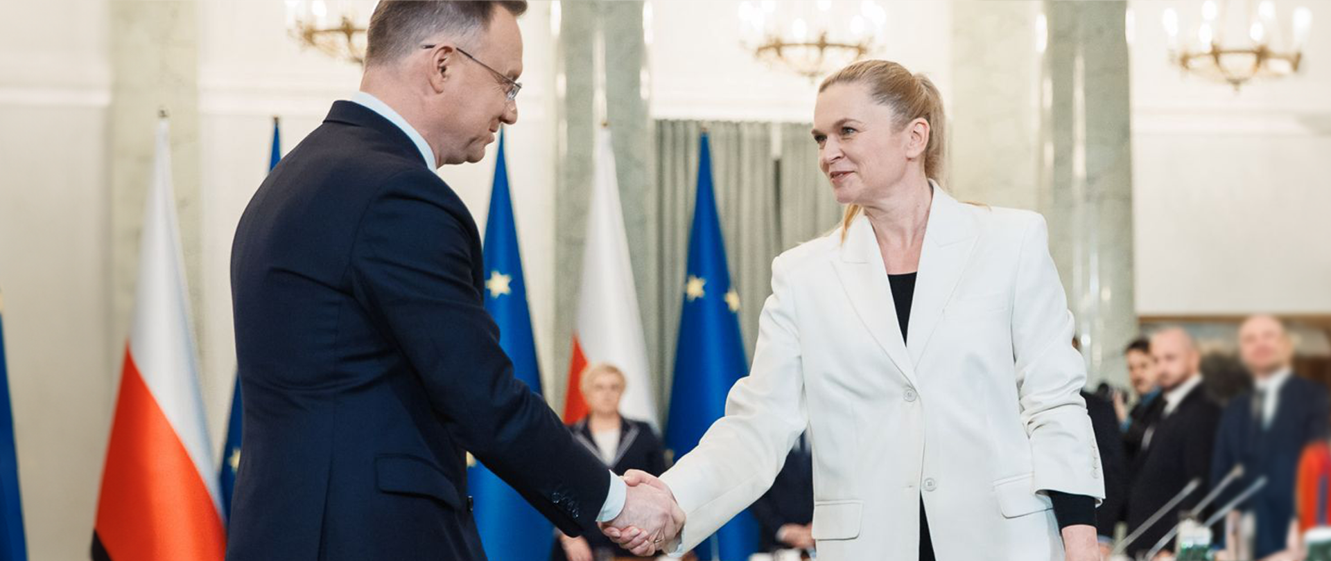 Minister Nowacka ubrana w białą marynarkę podaje rękę Prezydentowi Andrzejowi Dudzie. W tle widok sali, której obradowała Rada Gabinetowa.
