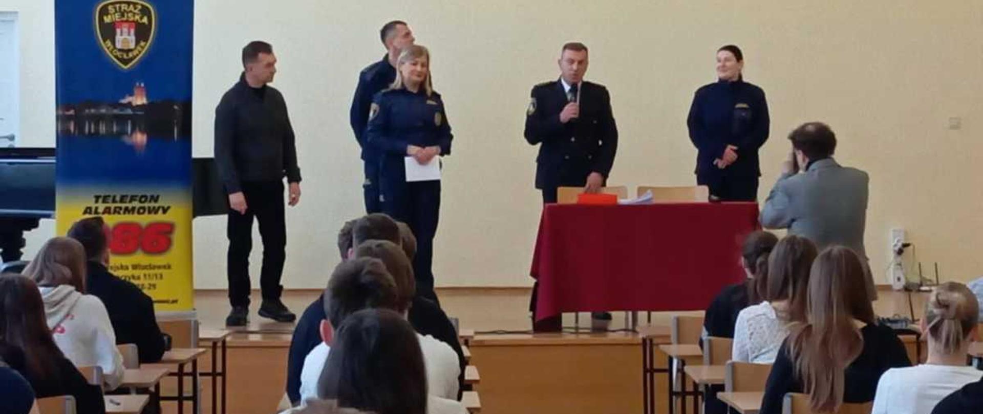 Zdjęcie przedstawia spotkanie edukacyjne zorganizowane w sali lekcyjnejw III Liceum Ogólnokształcącym im. Marii Konopnickiej we Włocławku (LMK), z udziałem funkcjonariuszy Straży Miejskiej we Włocławku.
Opis szczegółowy
Prelegenci: Przy stole, pod godłem Włocławka z napisem ""WŁOCŁAWEK LMK III"", stoi kilku funkcjonariuszy w mundurach. W centralnym punkcie mężczyzna w mundurze Straży Miejskiej przemawia do zebranych, trzymając coś w dłoniach. Towarzyszy mu dwóch innych umundurowanych strażników (jedna kobieta i jeden mężczyzna), a po lewej stronie stoi w cywilnym ubraniu (czarna bluza i spodnie) inny mężczyzna.
Publiczność: Na pierwszym planie i w środkowej części sali, tyłem do obiektywu, siedzą uczniowie w ławkach, słuchający prelekcji. Wśród nich, po prawej stronie, widać siedzącego mężczyznę w szarym garniturze.
Sala: Na ścianach wiszą plakaty informacyjne. Po lewej stronie widoczny jest plakat z logiem ""STRAŻ MIEJSKA"" i numerem alarmowym ""986"" oraz widokiem nocnego Włocławka. Po prawej, za stołem, znajduje się ekran lub tablica.
Atmosfera: Scena ukazuje formalne spotkanie, podczas którego przedstawiciele służb mundurowych przybliżają uczniom zasady bezpieczeństwa, co jest częścią działań profilaktycznych prowadzonych przez Straż Miejską we włocławskich szkołach.