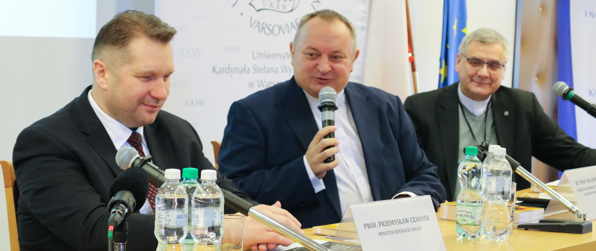 Minister Przemysław Czarnek siedzi przy stole podczas konferencji, przed sobą ma mikrofony, obok niego siedzą przedstawiciele UKSW w Warszawie.