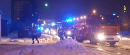 Na zdjęciu widać pojazdy straży pożarnej oraz ZRM z załączonymi sygnałami świetlnymi. Trzech ratowników znajdujący się w pobliżu pojazdów pożarniczych składa sprzęt użyty w działaniach gaśniczych przy pożarze piwnicy (węże, prądownice).