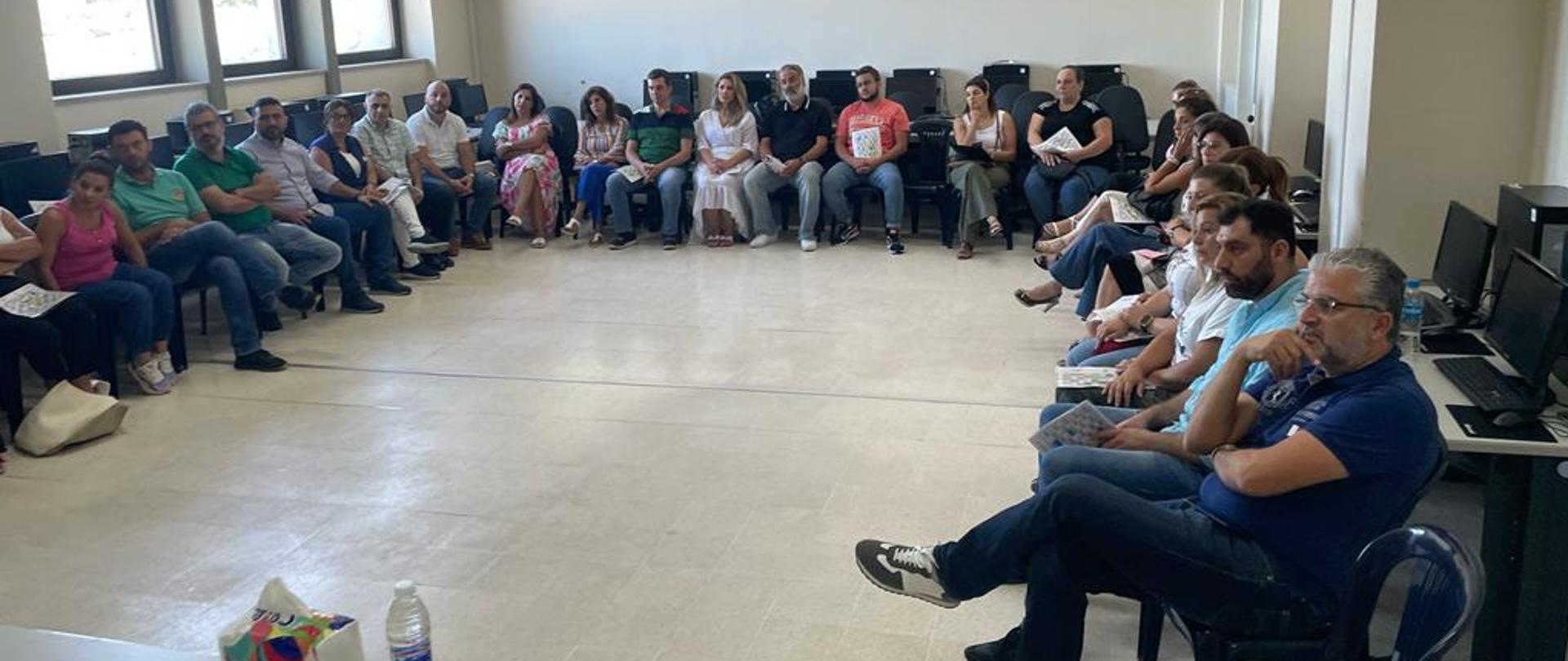 W sali na ustawionych pod ścianami i oknami krzesłach siedzi grupa dorosłych osób i patrzy się na prowadzącego szkolenie