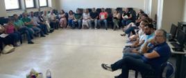 W sali na ustawionych pod ścianami i oknami krzesłach siedzi grupa dorosłych osób i patrzy się na prowadzącego szkolenie