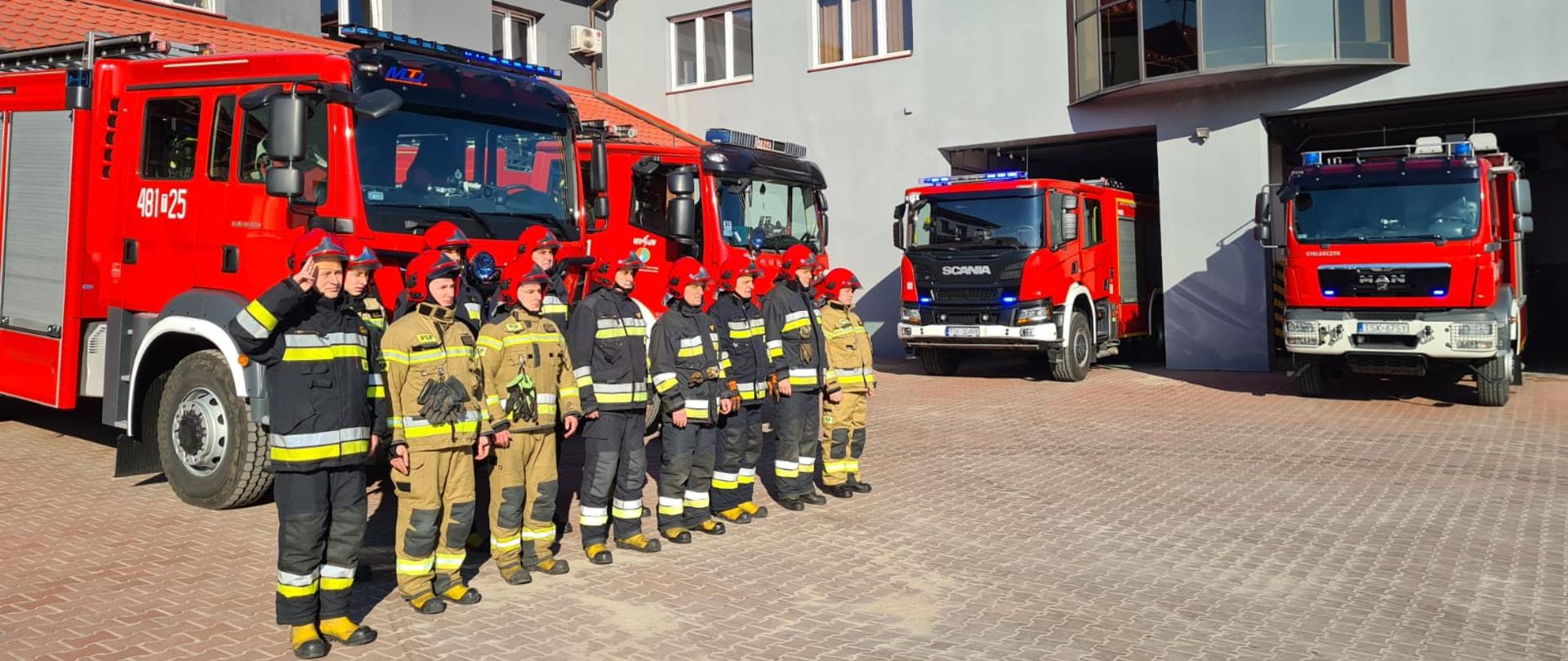 Zdjęcie obrazuje strażaków stojących na baczność, oddających honor strażakom z Ukrainy. Strażacy stoją w dwuszeregu przed samochodami pożarniczymi wystawionymi przed garaże KP PSP Skarżysko-Kamienna. W tle widać budynek komendy. 