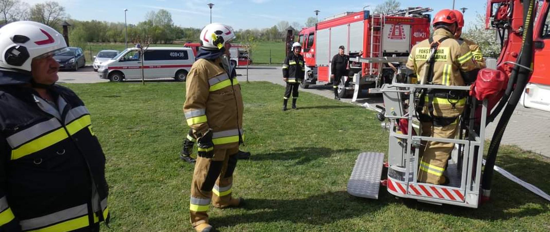 Po lewej stronie na trawie stoi dwóch strażaków Ochotniczej straży pożarnej ubranych w białe hełmy oraz ubrania koloru czarnego, po prawej stronie strażacy PSP w koszu podnośnika hydraulicznego. W tle samochód strażacki oraz 2 strażaków i drzewa 