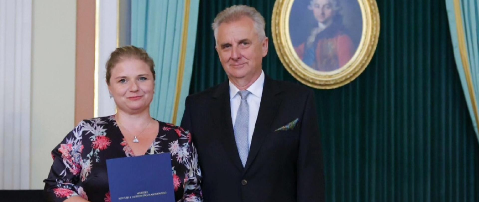 na zdjęciu na tle ciemnej ściany ze obrazem w złotej ramie kobieta i meżczyna elegancko ubrani kobieta trzyma w rękach niebieską teczkę