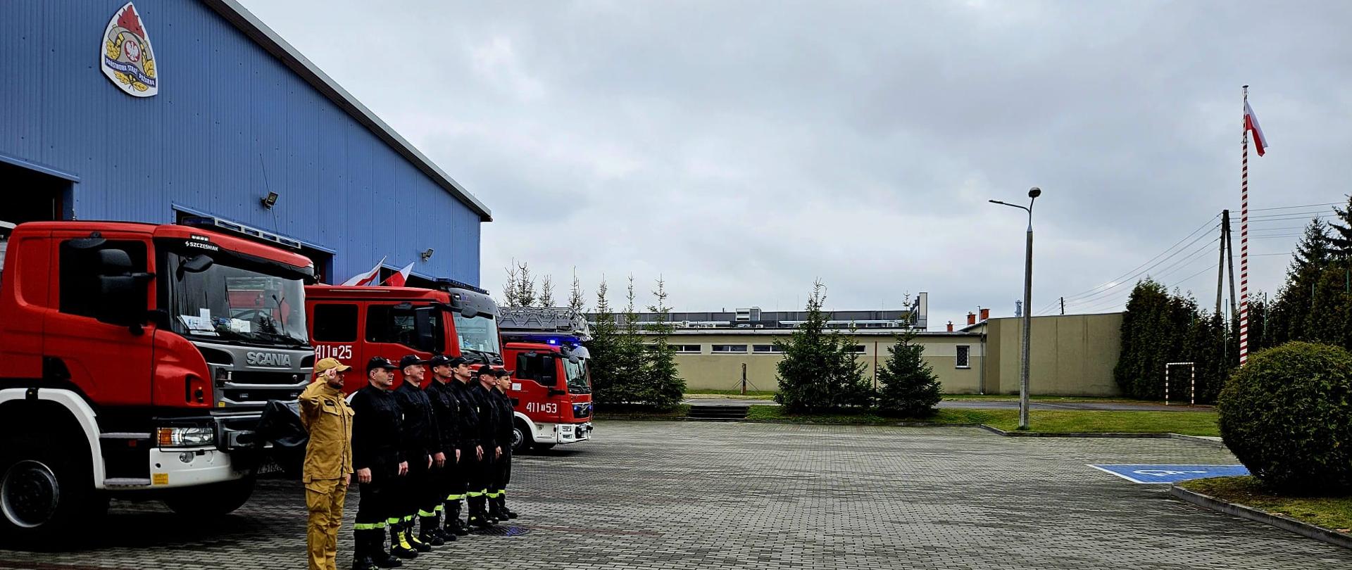 Zdjęcie obrazuje strażaków w umundurowaniu koszarowym. Strażacy znajdują się na tle samochodów ratowniczo-gaśniczych. Po prawej stronie widać maszt, na którym zawieszona jest biało-czerwona flaga.