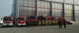 Na zdjęciu widzimy strażaków biorących udział w ćwiczeniach, stojących na tle 7 samochodów strażackich.