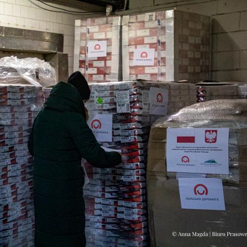 Zdjęcie przedstawia kobietę przyklejającą informację na palety z paczkami żywnościowymi 